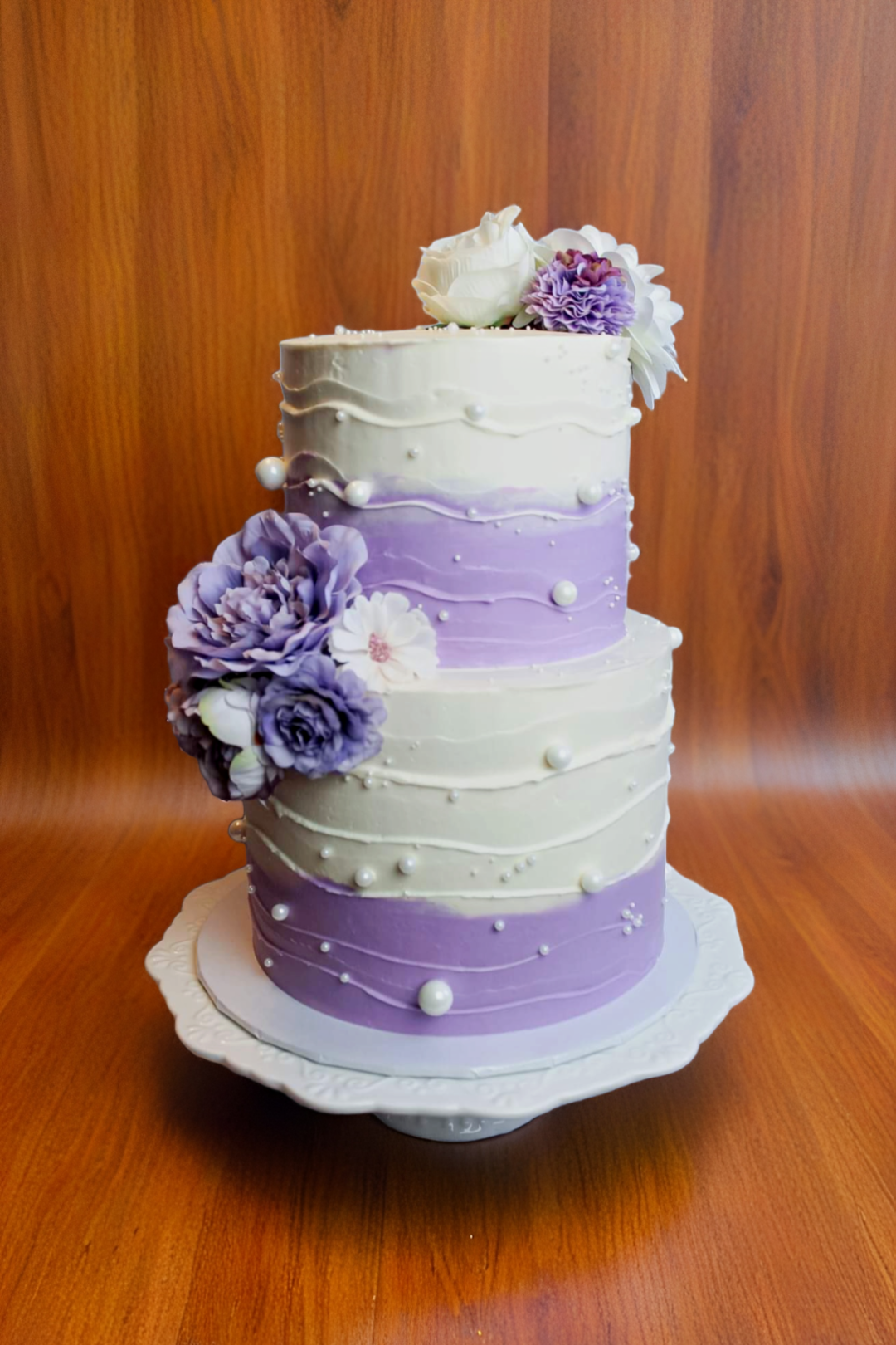 Two-tiered white and purple wedding cake with pearl details and floral topper
